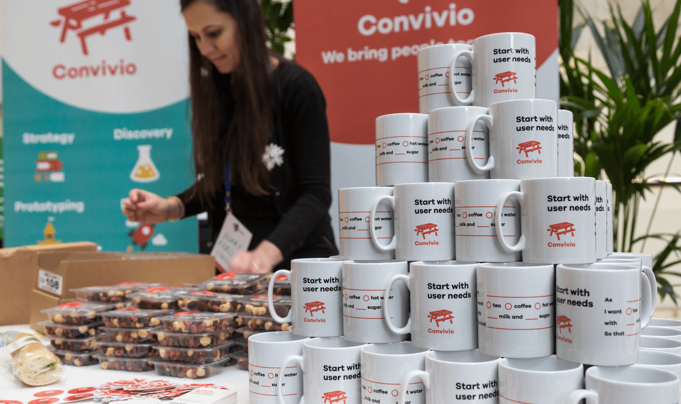 A photograph of a Convivio trade stand with a pile of mugs and banners in the background